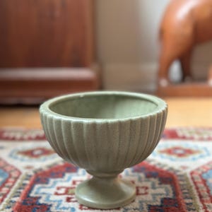 May include: A sage green, ribbed ceramic bowl with a pedestal base. The bowl has a rounded shape with vertical ridges and sits on a short, circular base. The bowl is displayed on a patterned rug with red, blue, and cream colors.