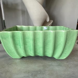 Peut inclure: Un pot de fleurs en céramique vert clair, de forme rectangulaire avec un bord festonné. Le pot présente des détails verticaux côtelés sur les côtés. Convient pour l'intérieur ou l'extérieur, c'est un objet décoratif.