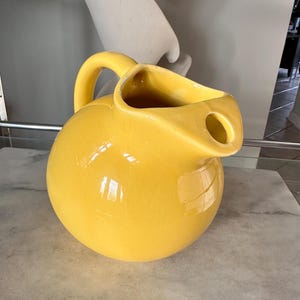 May include: A bright yellow ceramic pitcher with a rounded body and a curved handle. The pitcher has a spout with a circular opening. The glossy surface reflects light, highlighting its smooth texture. A decorative piece for serving beverages.
