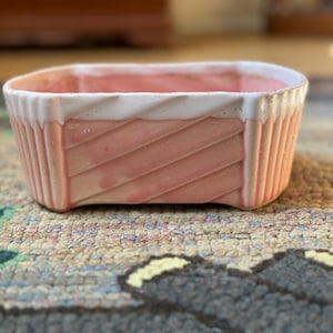 May include: A rectangular ceramic planter with a pink body and white rim. The planter features a diagonal ribbed design on the sides. The planter is sitting on a patterned rug.