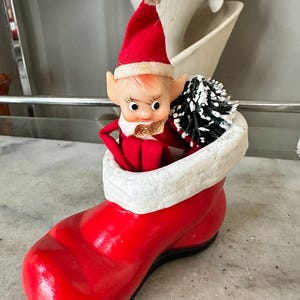 May include: A vintage Christmas decoration featuring a red boot with a white cuff, housing a small elf figure. The elf wears a red hat and outfit, with a small black and white tree. The boot is approximately 6 inches long.
