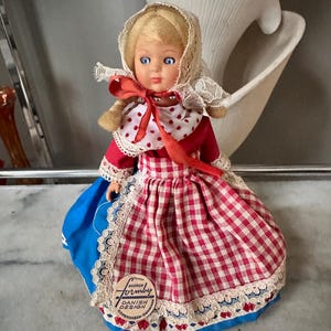 Puede incluir: Una muñeca vintage con vestimenta tradicional danesa. La muñeca tiene cabello rubio, ojos azules y viste un vestido a cuadros rojo y blanco con una falda azul. El atuendo de la muñeca incluye un gorro de encaje blanco y una cinta roja. La muñeca tiene una etiqueta que dice "Formby Danish Design Copenhagen Denmark."