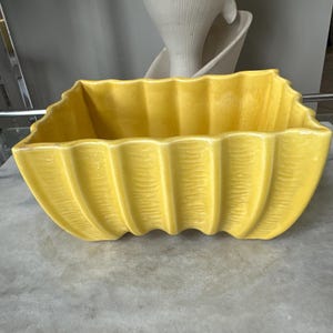 May include: A rectangular yellow ceramic planter featuring a scalloped, ribbed exterior design with vertical ridges. The vessel has a wide, open top and a continuous, wavy rim that follows the contours of the textured, glossy yellow surface.