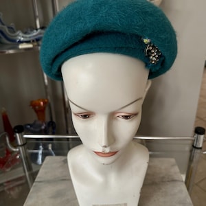May include: Teal-colored beret with a decorative jeweled embellishment. The hat is made of a soft, fuzzy material and is displayed on a white mannequin head. The beret has a classic, round shape and a slightly angled brim.