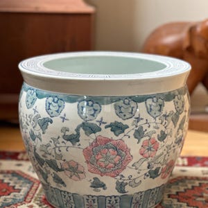 May include: A round ceramic planter featuring a white base with hand-painted floral motifs in shades of pink and muted blue. The rim is decorated with a geometric Greek key pattern, while the base displays a series of vertical rectangular panels.