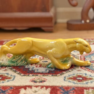 May include: A yellow ceramic figurine of a crouching panther with a glossy finish. The panther is depicted in a low, prowling stance with its head lowered and mouth slightly open. The base features green painted accents with gold detailing.