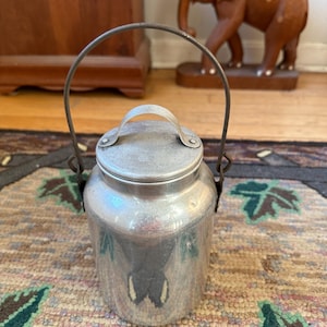 May include: A silver aluminum milk can with a wire handle and a lid. The can is sitting on a patterned rug.