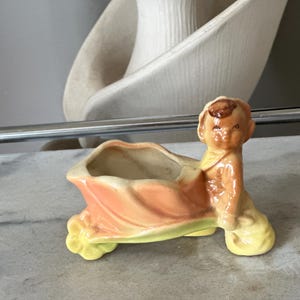 May include: A vintage ceramic planter in the shape of a baby. The planter is painted in shades of peach, yellow, and brown. The baby figure is seated, with a container for plants in front. A decorative piece.