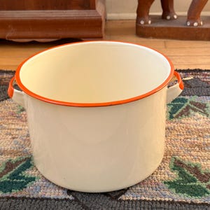 May include: Off-white enamelware pot with orange trim and handles. The pot is cylindrical with a wide opening and two small handles. The orange trim outlines the rim. The pot is sitting on a patterned rug.