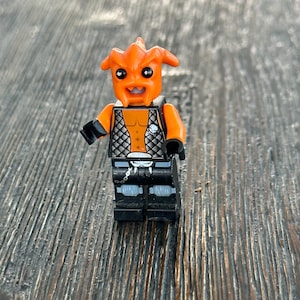 May include: An orange Lego minifigure with a horned head, black hands, and black boots. The torso features a silver vest over an orange chest. The figure is holding a small black accessory. The background is a wooden surface.