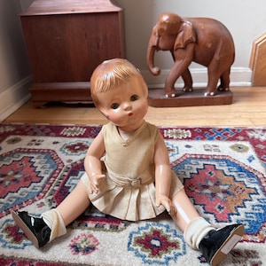 May include: A vintage doll with molded reddish-brown hair and green eyes, wearing a sleeveless tan top and a matching pleated skirt with a belt. The doll has light skin, wears white ribbed socks, and black patent leather shoes with bows.