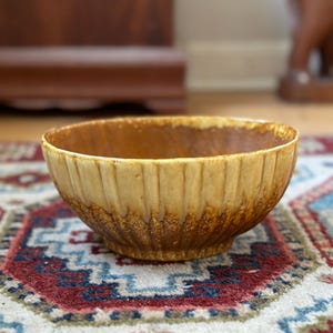May include: A ceramic bowl with a textured, ribbed exterior. The bowl has a warm, earthy colour palette, with shades of tan and brown. The interior is a lighter tan. The bowl is likely for serving or display.