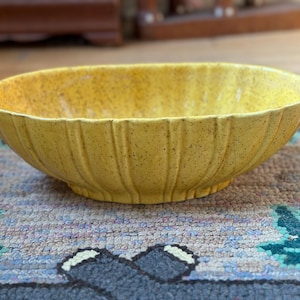 May include: A yellow ceramic bowl with a speckled finish. The bowl has a ribbed exterior and a wide, oval shape. The bowl is sitting on a patterned rug. The bowl is likely for serving food or as a decorative piece.