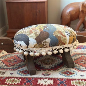 May include: A turtle-shaped footstool with a dark brown wooden head, tail, and four legs. The oval seat is upholstered in a fabric featuring a multi-colored duck and wheat pattern, finished with a cream-colored fringe trim with small pom-poms.