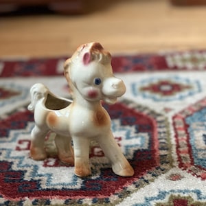 May include: A ceramic horse figurine with a planter opening. The horse is primarily white with brown spots and accents, including its mane, tail, and hooves. It has blue eyes and pink cheeks. The figurine is standing on a patterned rug.