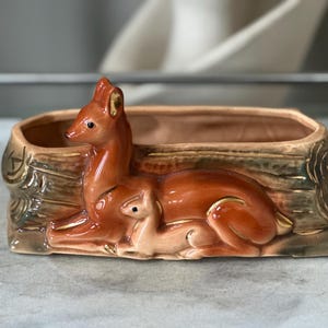 May include: A rectangular ceramic planter featuring a relief sculpture of a doe and a fawn resting against a textured log. The deer are finished in a glossy reddish-brown glaze with gold accents, while the log base has a muted brown and green finish.