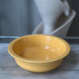 May include: A bright yellow ceramic bowl with a smooth, glossy finish and a wide, flat rim. The bowl features a series of concentric circular ridges along the exterior side, providing a subtle textured detail to the otherwise simple, round design.