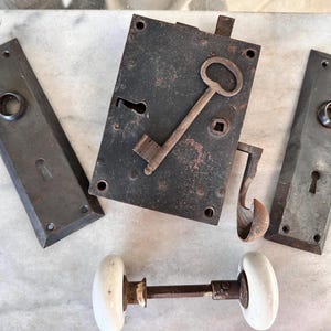 May include: An assortment of antique door hardware, including a lock plate with a key, two rectangular plates, and a doorknob with white ceramic knobs. The metal components have a weathered, dark finish, suggesting age and use.