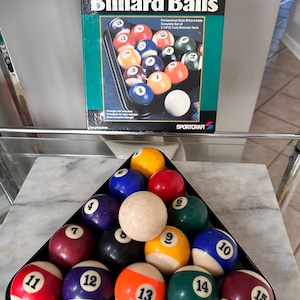 May include: A full set of numbered and colored billiard balls in a black triangle rack, with a white cue ball. The box behind the balls reads "Billiard Balls" and "SPORTCRAFT". The balls are 5.7cm in diameter.
