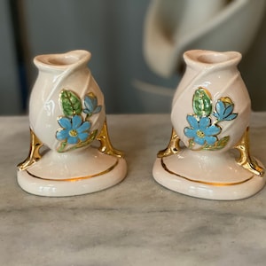 Puede incluir: Un par de candelabros de cerámica vintage. Cada soporte es de color blanco roto con una base y detalles dorados. Diseños florales azules con hojas verdes decoran la parte delantera de cada soporte. Los candelabros tienen una forma única y elegante.