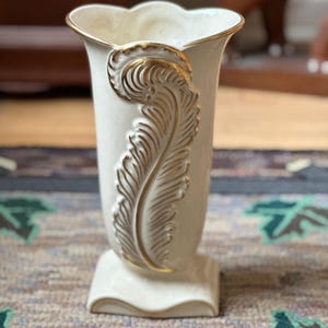May include: Cream-colored vase with a gold feather design and gold trim. The vase has a square base and a scalloped rim. The feather design is on the front of the vase. The vase is likely ceramic or porcelain.