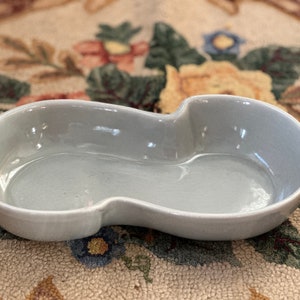 Puede incluir: Un plato de cerámica gris claro en forma de cacahuete. El plato tiene un acabado liso y brillante y un interior poco profundo. El diseño presenta dos secciones ovaladas conectadas, creando una pieza de servicio única. Ideal para servir aperitivos.
