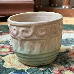 May include: A light green and brown ceramic planter with a decorative design. The planter has a scalloped rim and a textured surface.