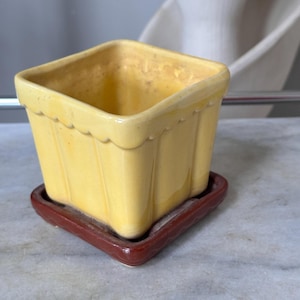 May include: A square, yellow ceramic planter with a scalloped rim, sitting on a rectangular, dark red ceramic saucer. The planter has vertical ribbed detailing. A decorative item for indoor plants.