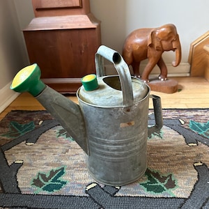 May include: A galvanized metal watering can with a green and yellow spout and handle. The watering can is sitting on a patterned rug. A wooden elephant statue is in the background.