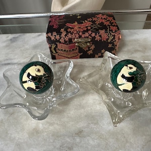 May include: Two spherical green cloisonné balls featuring a panda design, each resting in a clear glass star-shaped dish. Behind the dishes sits a rectangular black fabric box decorated with a red and gold floral pattern and a metal latch.