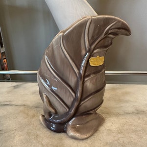 May include: A ceramic leaf-shaped vase in shades of gray and brown. The vase has a textured surface with detailed veining and a small gold label. The base is a rounded shape, and the overall design is reminiscent of nature.