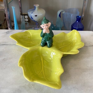 May include: A green ceramic leaf-shaped serving dish with three sections. A small green ceramic elf figurine sits in the centre section.