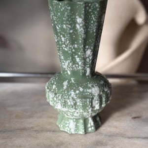 May include: A green ceramic vase with a speckled white pattern. The vase has a fluted, angular body and a rounded base. The vase is approximately 6 inches tall.