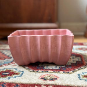 May include: A square, pale pink ceramic planter with a fluted design. The planter is placed on a patterned rug with red, blue, and cream colours. A wooden cabinet and a light-coloured wall are in the background.