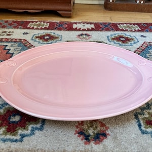 May include: A large, oval pink serving platter with decorative handles. The platter has a glossy finish and is set on a patterned rug. The handles feature a scroll design.
