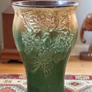 May include: A tall, ceramic vase with a glossy green and tan finish. The vase features embossed floral designs and a dark brown rim. The vase has a flared top and a gently curved body, suitable for indoor decor.