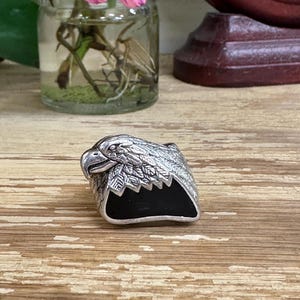May include: A silver-toned ring featuring an eagle head design with intricate feather detailing and a black enamel base. The ring is set against a wooden surface, with a glass jar of flowers in the background.