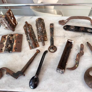 May include: A collection of antique metal hardware pieces, including hinges, latches, handles, and decorative elements. The items display a weathered, rusty appearance with remnants of paint, suggesting age and use. The hardware is arranged on a light-colored surface.