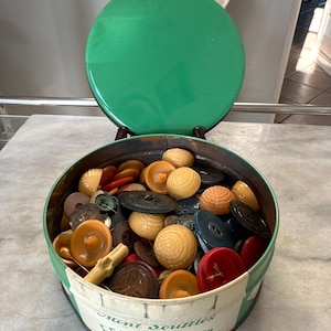 May include: A round metal tin with a green lid and a cream-colored base featuring the text E.G. WHITMAN & CO. PHILADELPHIA, PA. The tin is filled with a collection of assorted vintage buttons in various colors, shapes, and textures, including round, flat, and shank styles.