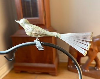 May include: A glass bird ornament with a white iridescent finish and a long tail of white fibres with gold tips. The bird is perched on a black metal stand.
