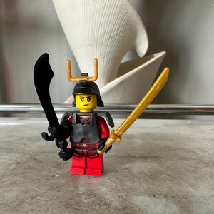 Pode incluir: Minifigura Lego de um guerreiro, vestida com armadura vermelha e preta, segurando uma espada preta curva na mão esquerda e uma espada dourada na direita. A figura tem uma cabeça amarela com um capacete preto e detalhes dourados.