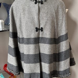 May include: A gray and charcoal striped cape with a textured weave. The cape features a decorative collar and trim, and is fastened with two black clasps. The cape is displayed on a mannequin.