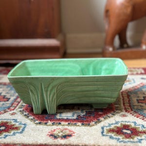 May include: A rectangular, light green ceramic planter with a textured surface and stylized Art Deco design. The planter sits on four small feet and is displayed on a patterned rug.