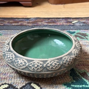 May include: A green glazed ceramic bowl with a decorative, embossed pattern around the rim. The bowl is sitting on a patterned rug.