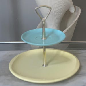 May include: Two-tiered serving tray with a silver handle. The top tier is a light blue, and the bottom tier is a pale yellow. The tray is designed for serving desserts or appetizers.