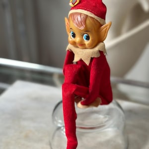 May include: A vintage Christmas elf doll with a red felt outfit and hat, tan skin, and blue eyes. The elf is sitting on a clear glass jar. The elf's hat has a gold trim.