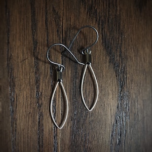 May include: A pair of silver-colored, open teardrop earrings with dark gray accents. The earrings hang from a curved hook. The jewelry is displayed on a dark wood surface.