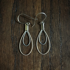 May include: A pair of silver-toned teardrop earrings. Each earring features a double teardrop design, with a smaller teardrop nested inside a larger one. The earrings hang from a simple hook. The background is a dark wood surface.