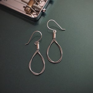 May include: Silver-toned teardrop earrings with a minimalist design. The earrings feature a simple wire loop and a hook closure. The background includes a portion of a silver instrument, set against a dark green surface.