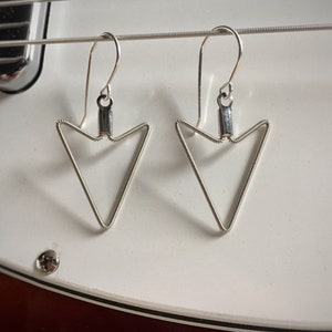 May include: A pair of silver triangle shaped earrings with a wire design. The earrings are hanging from a silver hook.